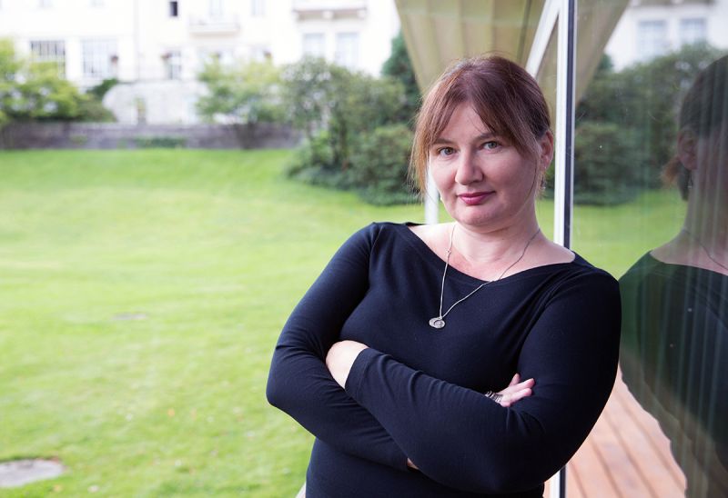 Color photograph of the head and torso of a light skinned woman with brown hair and wearing a black long sleeved top and a necklace; she crosses her arms as she leans against an exterior glass window of a building with a green grass yard