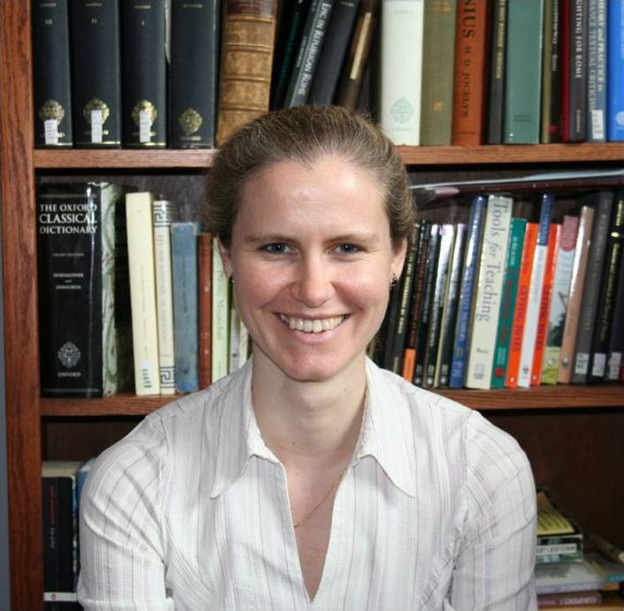 Color photograph of the head and shoulders of a light skinned woman wearing a white shirt and smiling at the camera; she sits in front of a wooden bookcase with books