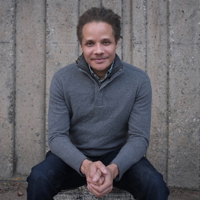 Color photograph of a brown skinned man wearing a gray sweater and blue jeans; he sits on front of a concrete wall and smiles slightly at the camera
