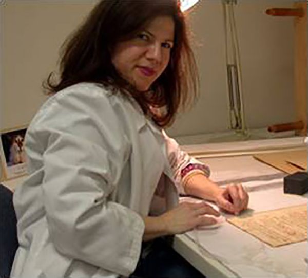 Color photograph of light skinned woman wearing a white lab coat; she is seated at a work table and looks up at the camera