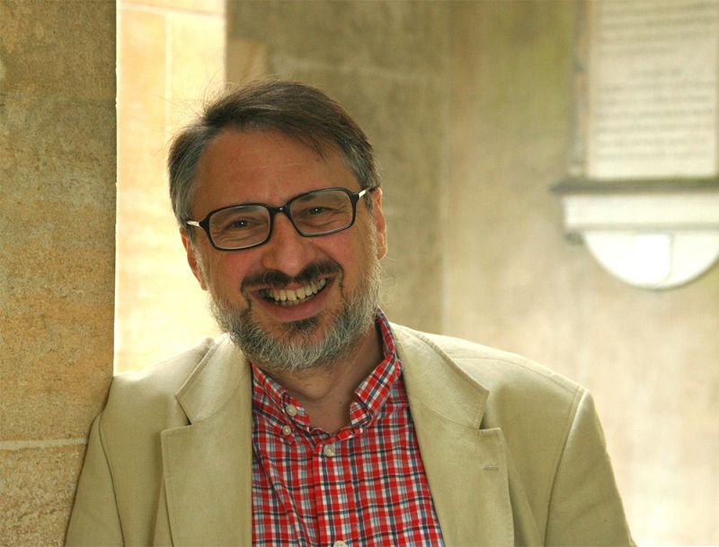 Color photograph of a light skinned man wearing a light tan blazer and red button up shirt leaning against a wall; he smiles at the camera