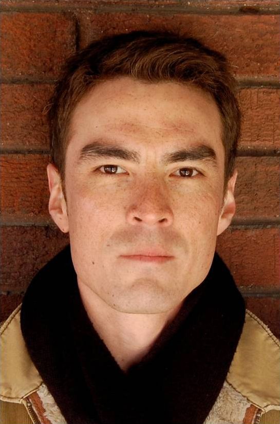 Color photograph of the face of a light skinned man standing against a brick wall and wearing a black scarf