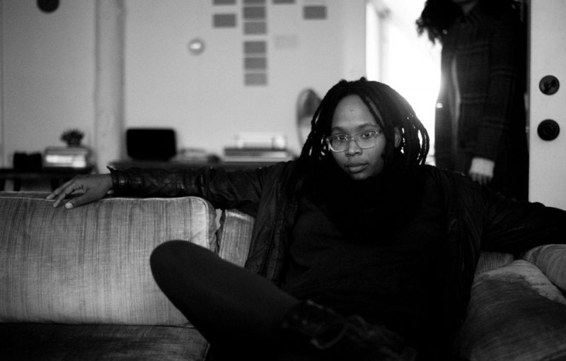 Black and white portrait of Jennifer Packer seated on a sofa, wearing glasses with a crossed leg and an arm stretched across the back of the sofa