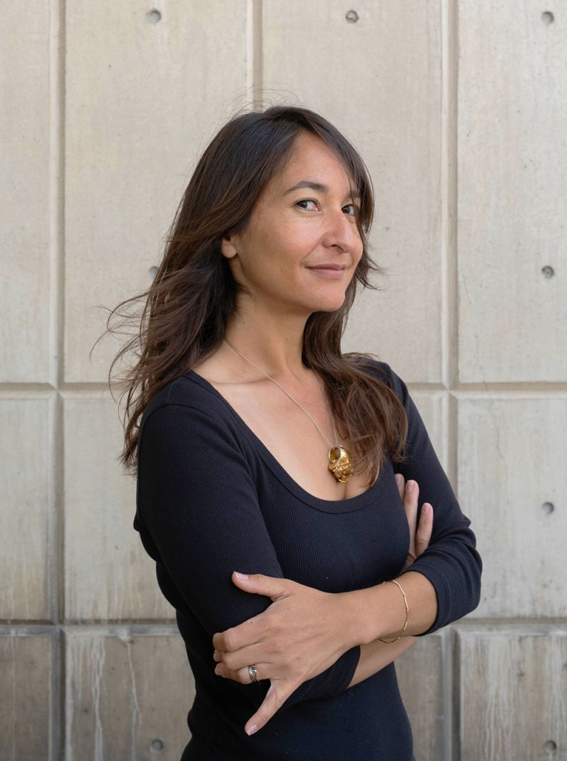 Color photo of an Asian woman with dark hair standing against a concrete wall and smiling at the camera