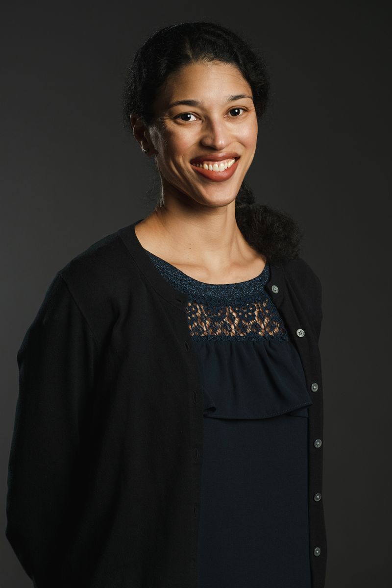 Color photograph of a brown skinned woman in a black dress, smiling widely at the camera