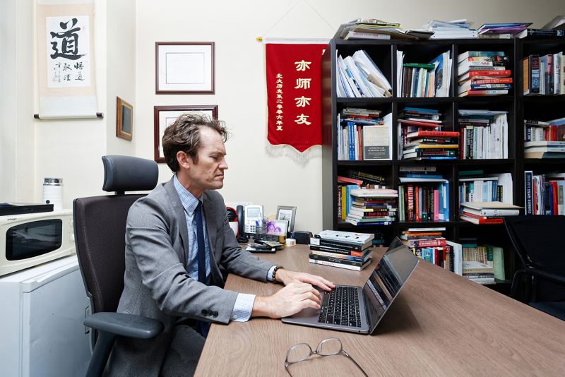 Color photograph of a light skinned man wearing a blazer and sitting at a desk in a book-lined professor's office, typing on a laptop computer