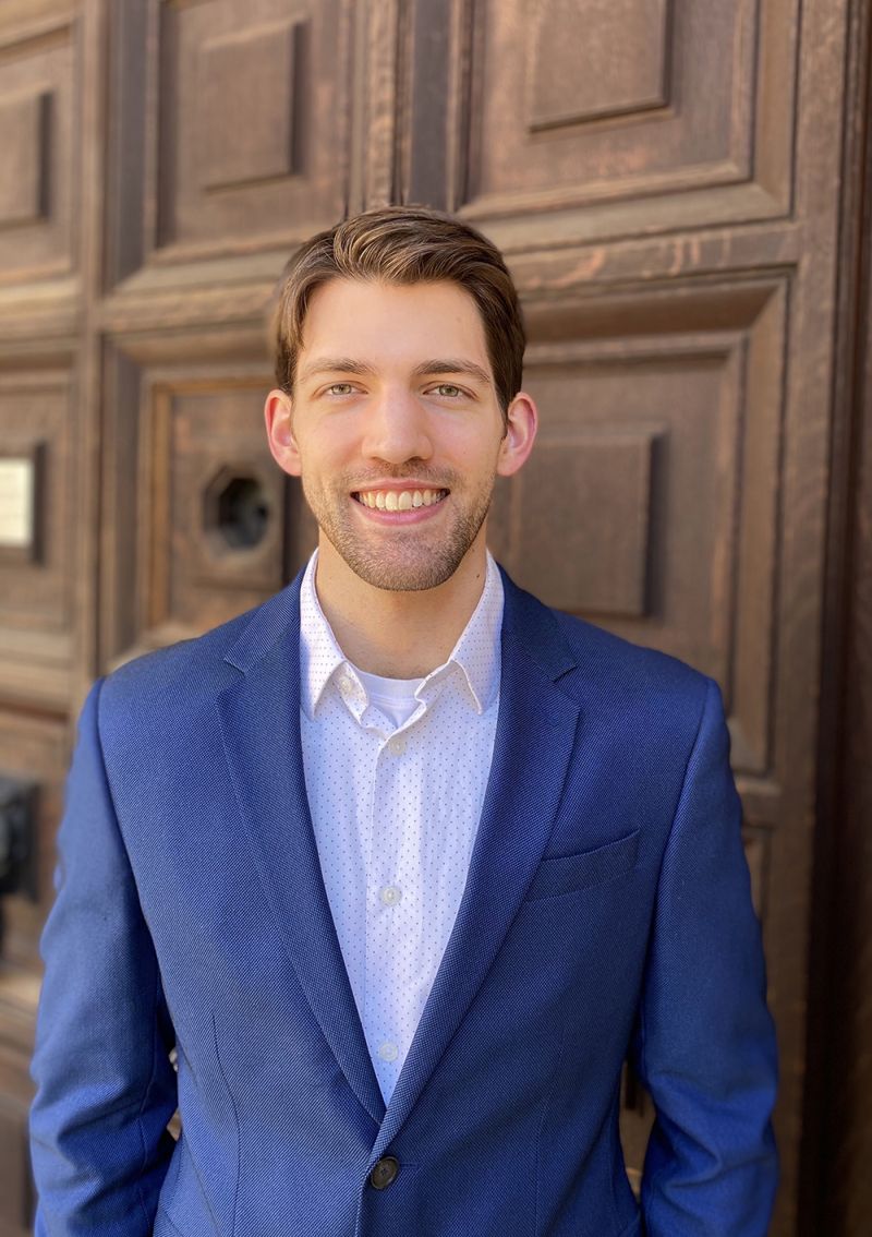 Color portrait of the head and torso of John Izzo, standing in front of a large wooden door and wearing a blue suit