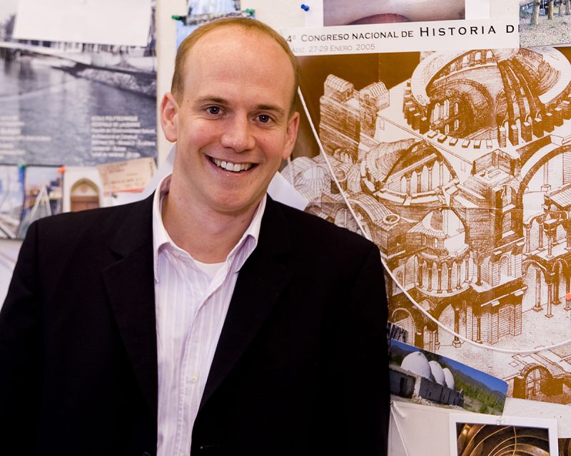 Color photograph of a light skinned man wearing a shirt and dark blazer and smiling at the camera
