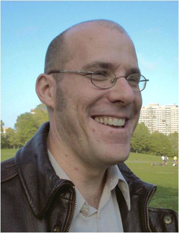 Color photo of the head and shoulders of a light skinned man wearing glasses and standing in a park