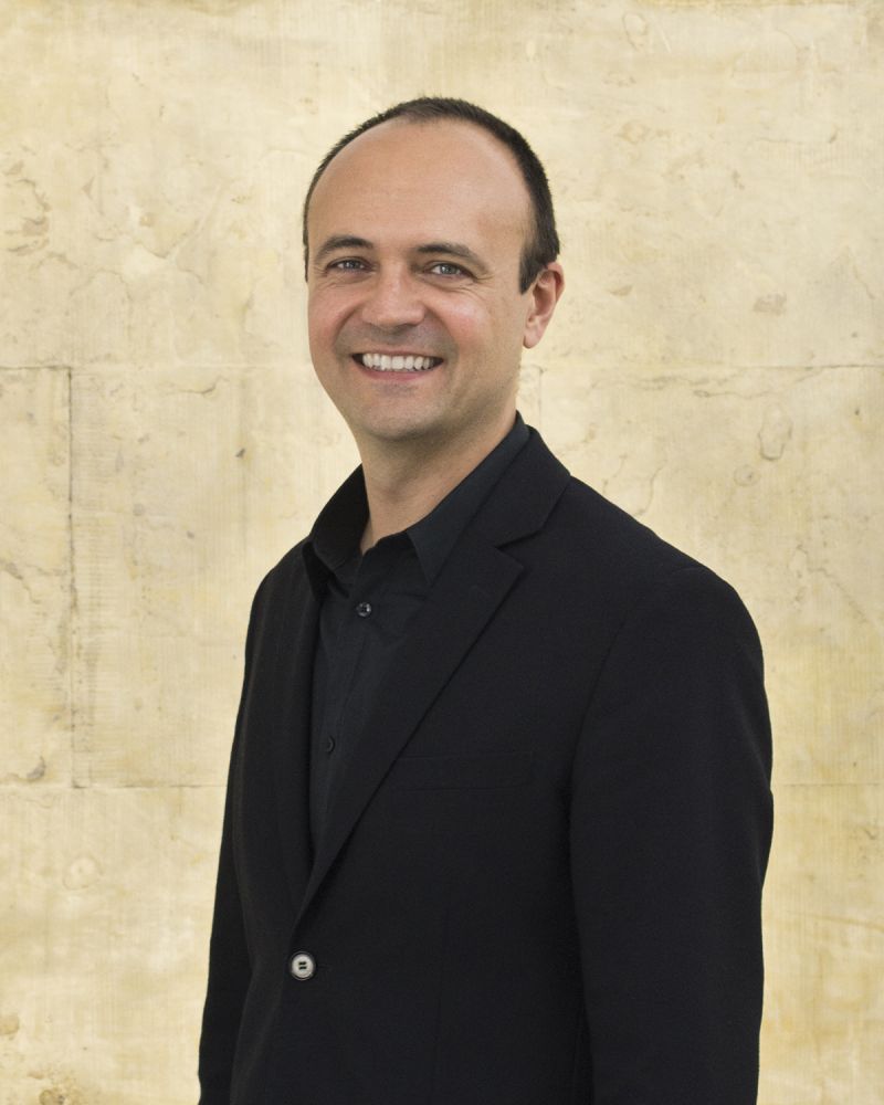 Color photograph of the Jorge Otero-Pailos from the waist up; he is light skinned, has closely cropped fair, wears a black shirt and blazer, and smiles directly at the camera