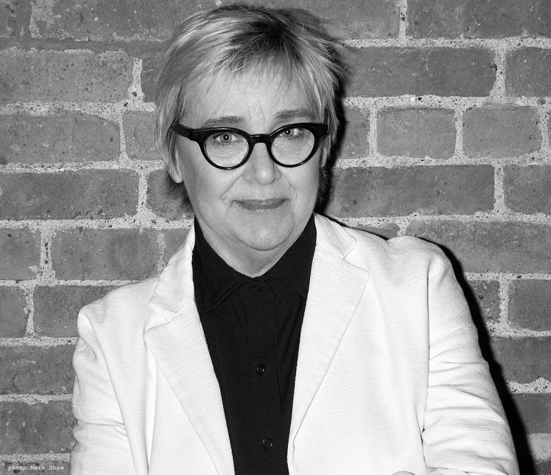 Black and white portrait of a light skinned woman wearing glasses and looking at the camera in front of a brick wall
