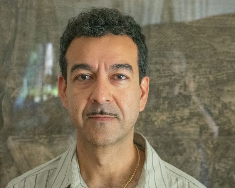 Color portrait of the head and shoulders of an olive skinned man with a thin mustache looking at the camera