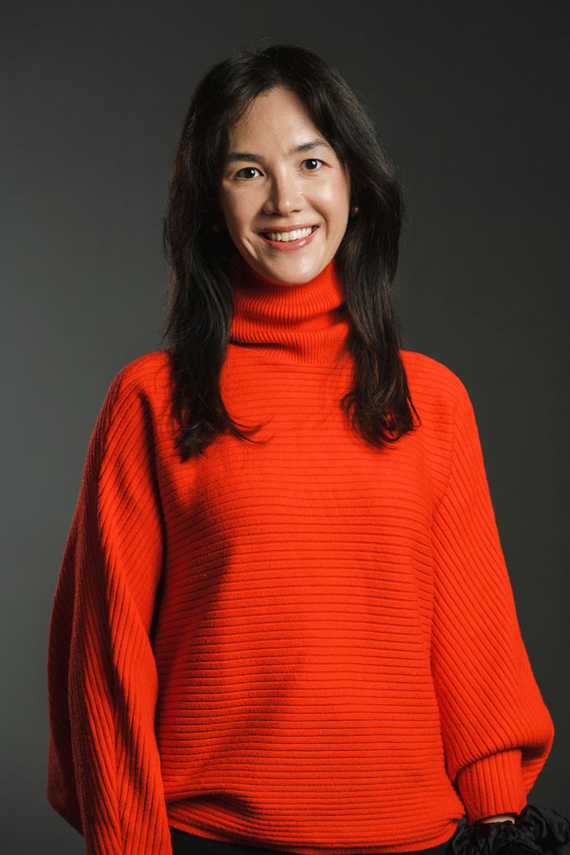 Color photograph of an olive skinned woman with dark hair wearing a bright orange dress in a photographer's studio, smiling at the camera