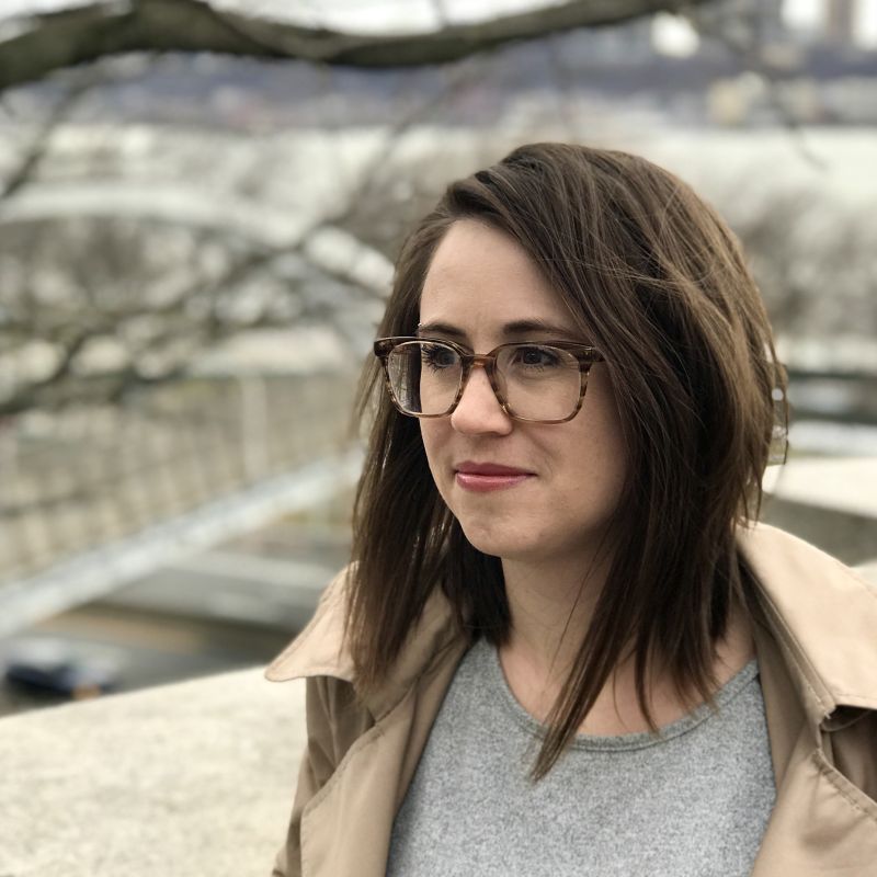 Color portrait of Katherine Balch, turned three-quarters, wearing glasses and a tan coat over a gray top, and standing next to a concrete ledge in an outdoor location