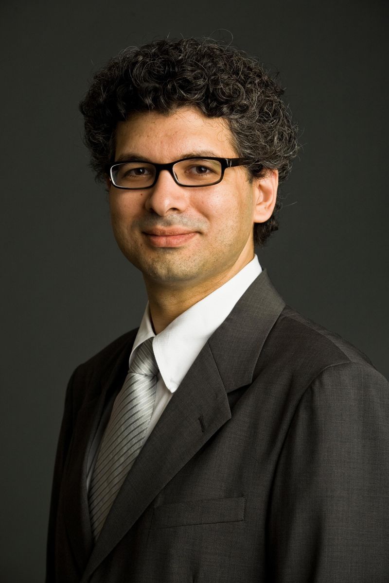Color photograph of a light skinned man wearing glasses, a suit, and a tie in a photographer's studio, smiling at the camera
