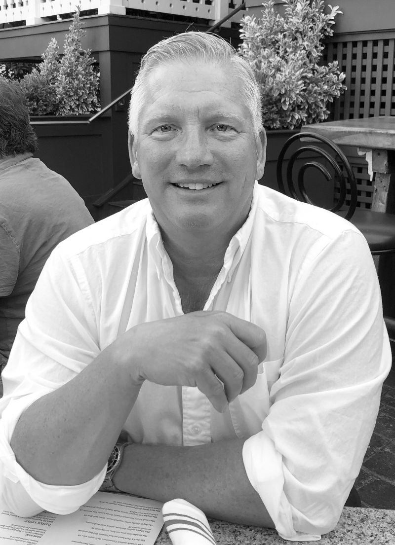 Black and white portrait of the torso and arms of Kevin Benham, seated at an outdoor table