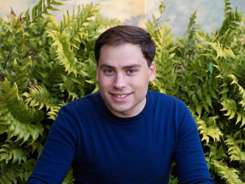 Color photograph of Kevin Ennis, wearing a blue sweater and sitting in an outdoor location with green plants behind him