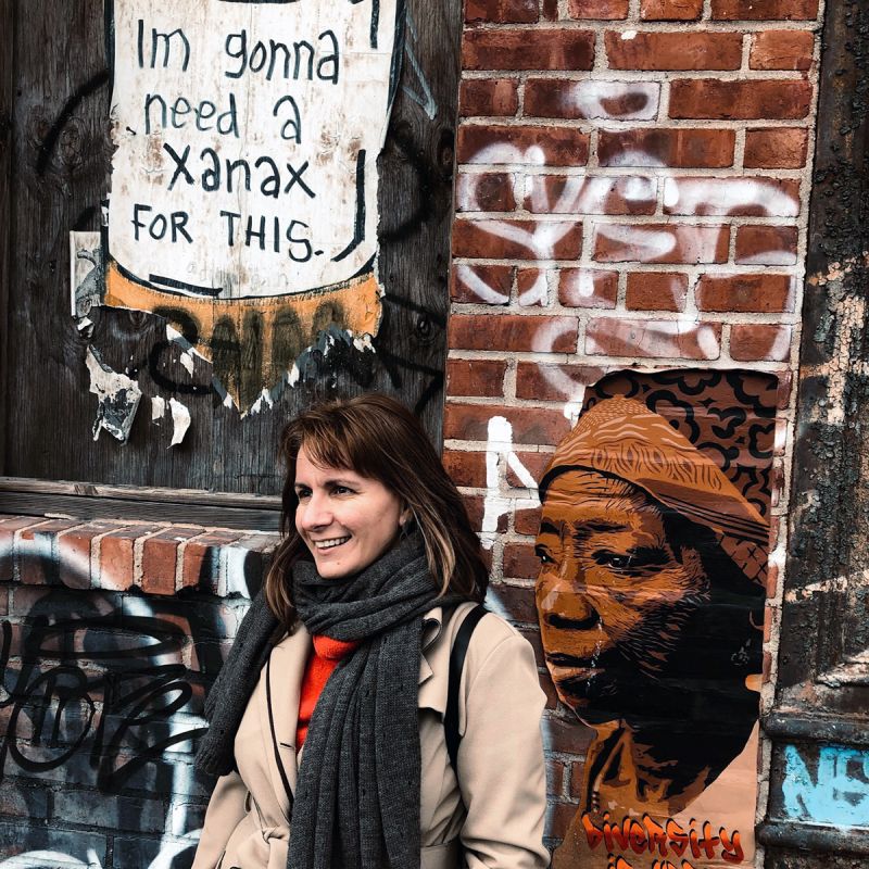 Color photograph of the head and torso of a light skinned woman with dark hair and dressed in a tan coat and scarf; she stands in front of a brick wall with much graffiti and looks off camera
