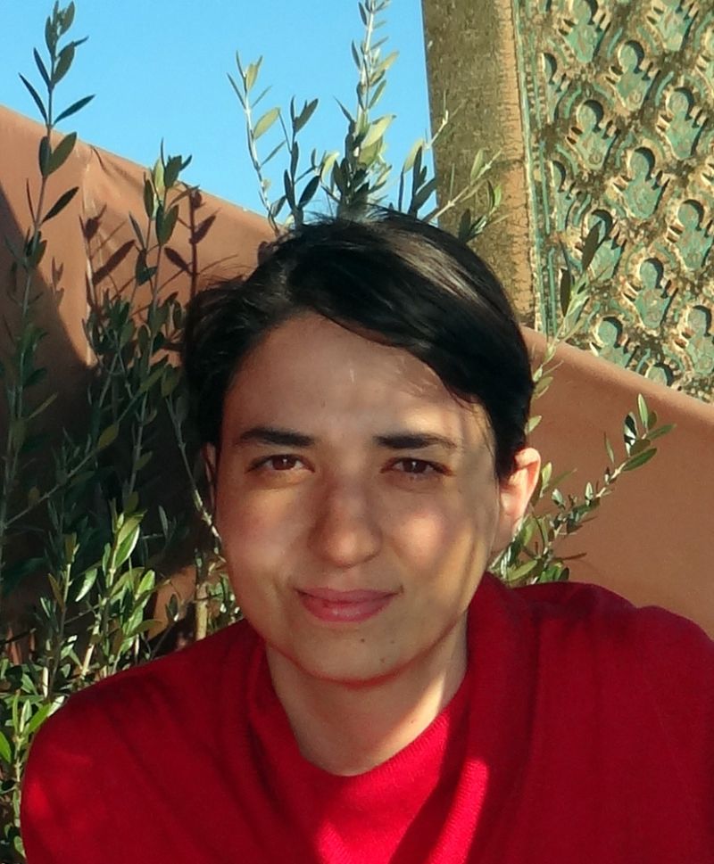 Color photograph of the head and shoulders of a light skinned woman wearing a red shirt and sitting in front on a plant and two walls, one slanted and the other patterned