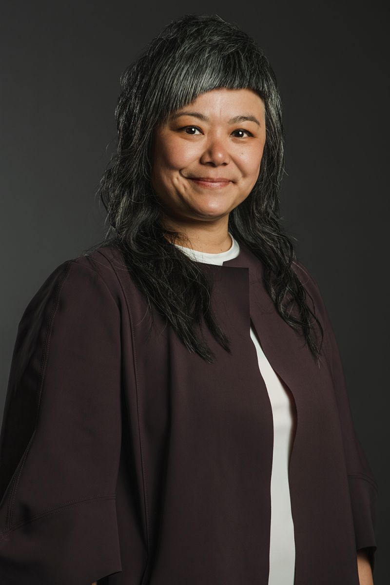 Color photograph of an Asian woman with black and gray hair and a black jacket; she smiles at the camera