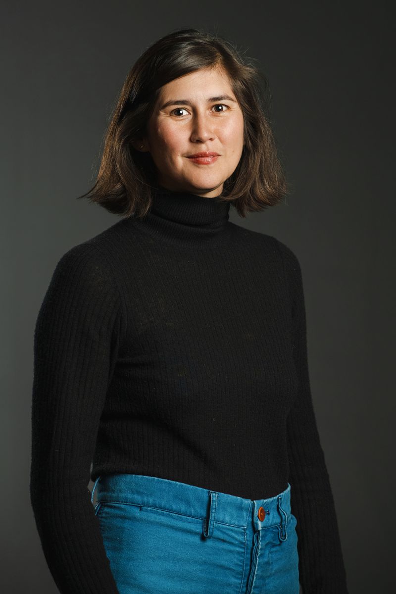 Color photograph of a light skinned woman with dark hair wearing a black turtleneck and blue jeans, looking at the camera