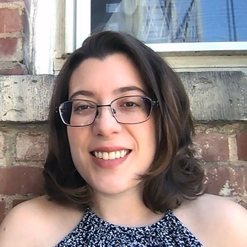 Color photograph of the head and shoulders of a light skinned woman with brown hair smiling at the camera; she stands against a red brick wall that also contains the bottom corner of a window and cement windowledge