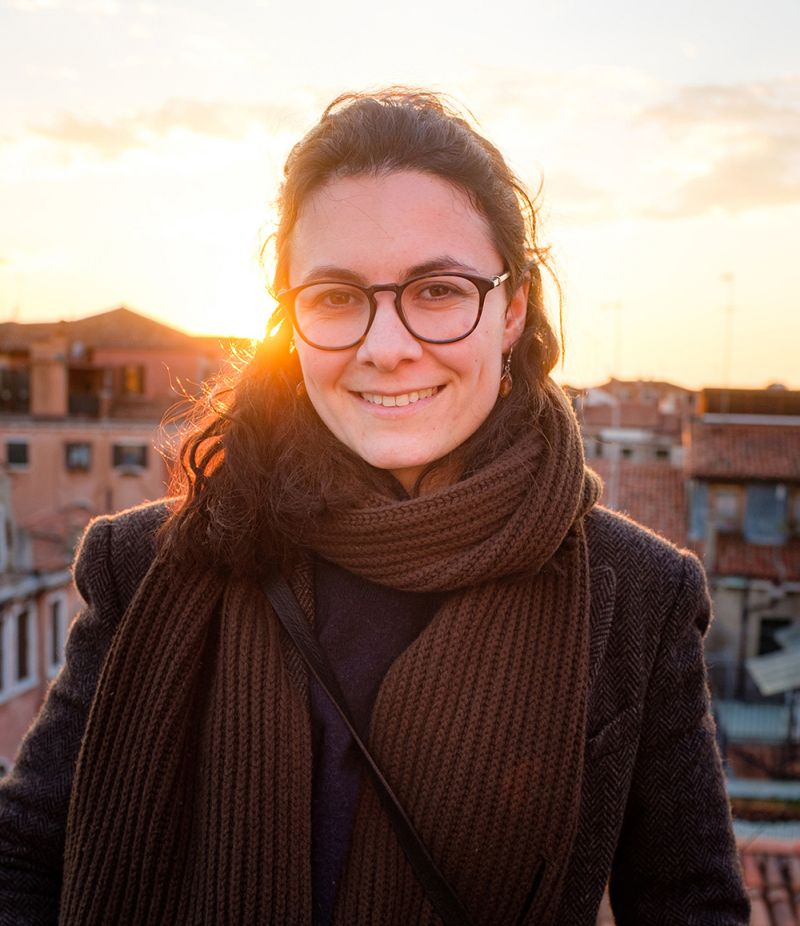 Color photograph of the head and torso of Lillian Datchev standing on an Italian rooftop or balcony, with the sun setting behind her