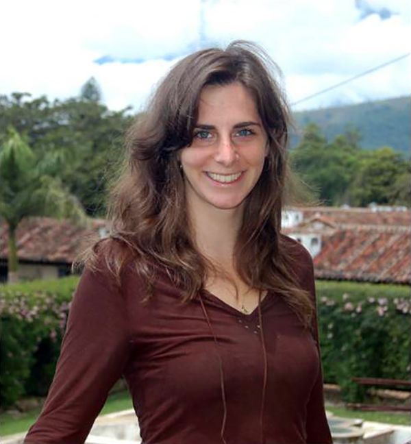 Color photograph of a light skinned woman with brown hair seen from the waist up; she is standing outdoors (with hills and roofs behind her) and smiles at the camera