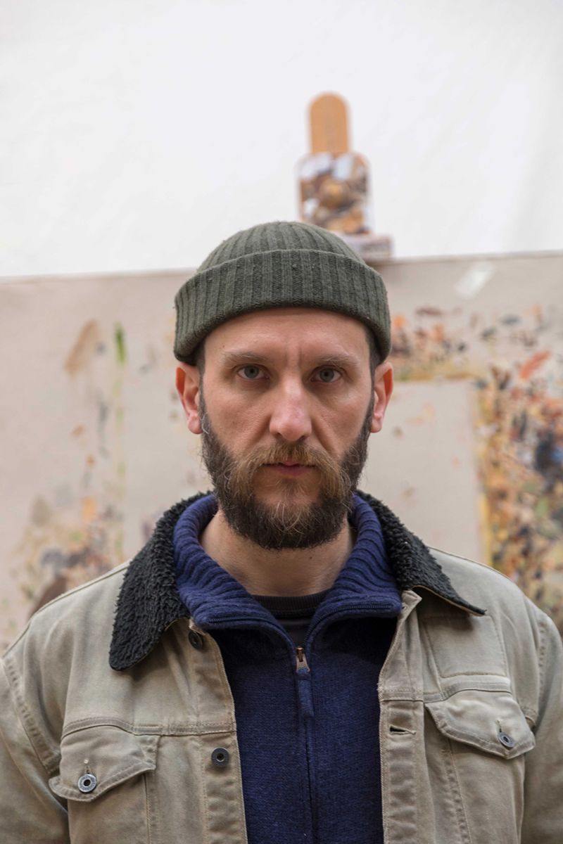 Color photograph of the head and torso of Manuele Cerutti standing in front of a painting easel