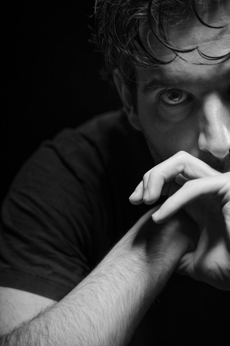 Black and white photograph showing the cropped head, shoulder, forearm, and hand of a light skinned man with dark hair, who looks intensely at the camera