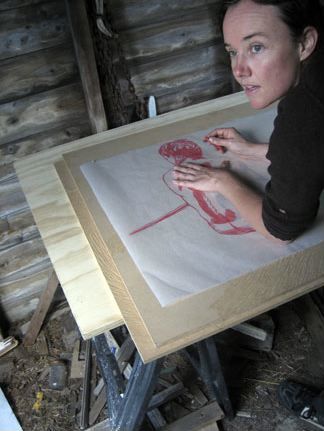 Color photograph of the head and shoulder of a light skinned woman working at a drafting table, with a drawing in front of her; she looks to the left offscreen