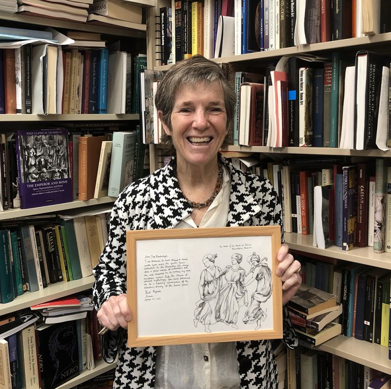 Color photograph of Mary Tolly Boatwright standing in front of book-filled shelves and holding a framed handwritten letter and drawing