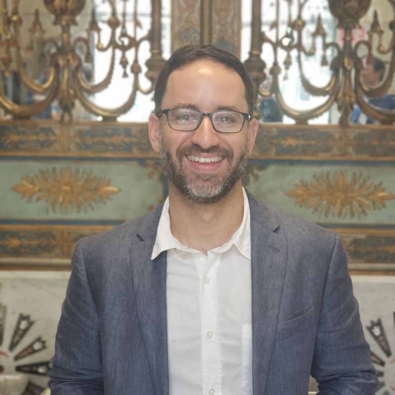 Color portrait of the head and torso of Matthew H. Ellis, facing forward, wearing glasses, and standing in front of an ornately decorated wall with a mirror