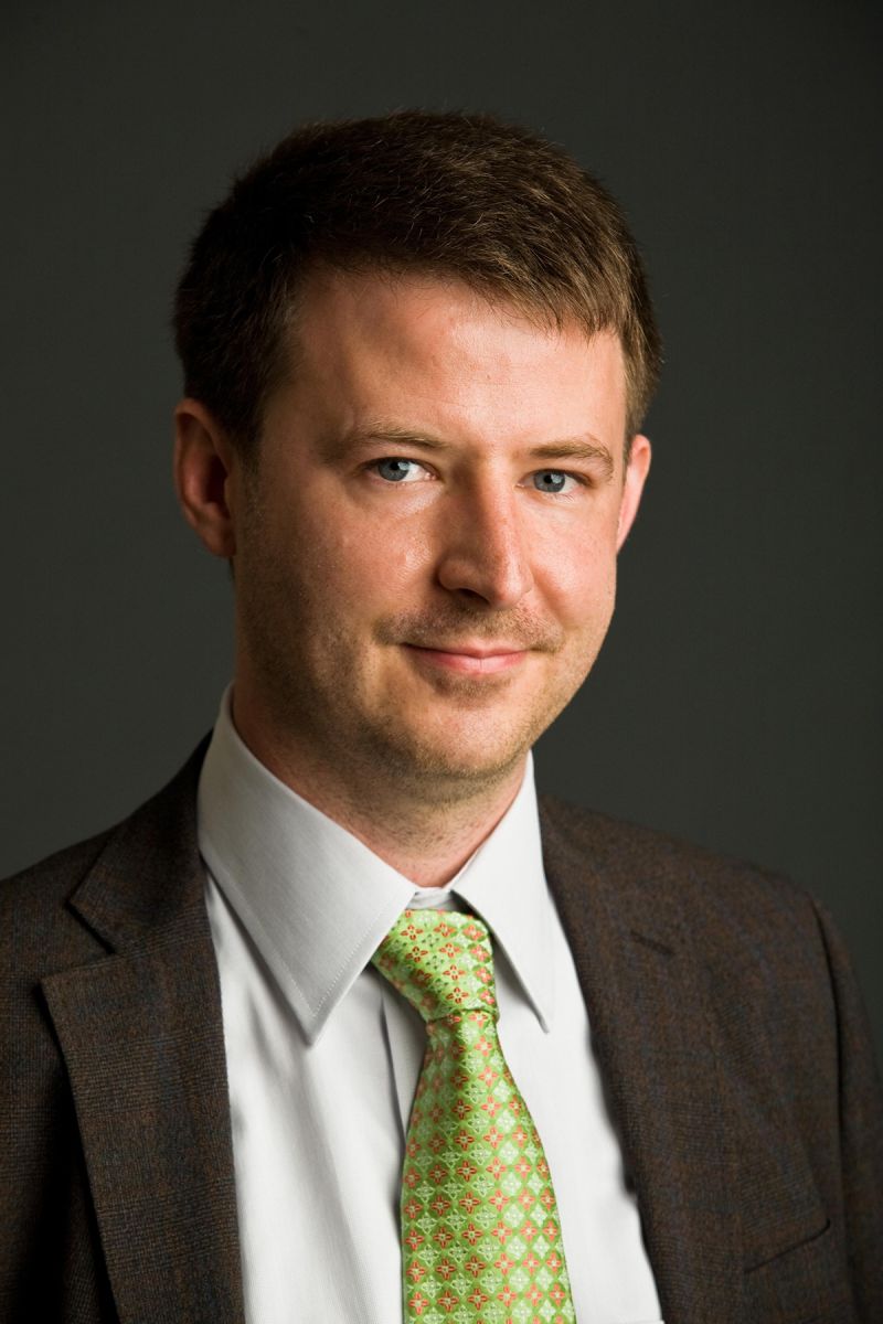 Color photograph of the head and torso of a light skinned man in a photographer's studio wearing a dark suit and green tie and smiling at the camera
