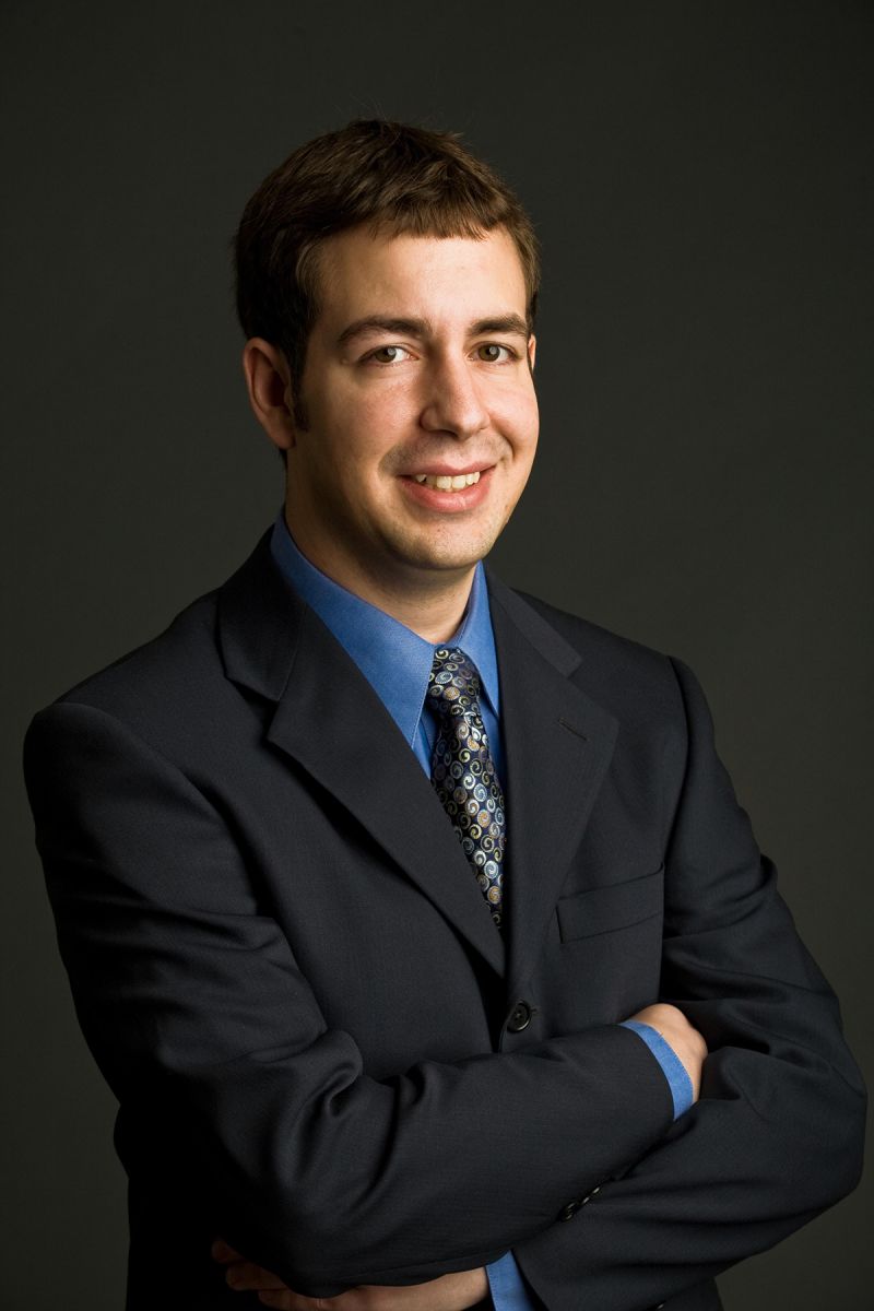Color photograph of the head and torso of a light skinned man in a photographer's studio wearing a suit and smiling at the camera