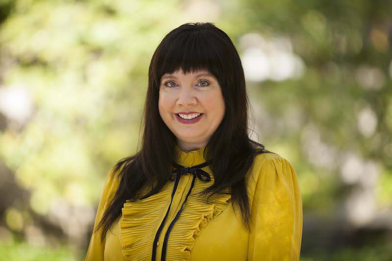 Color photograph of the head and torso of Melanie Masterton Sherazi, standing outside and wearing a yellow dress
