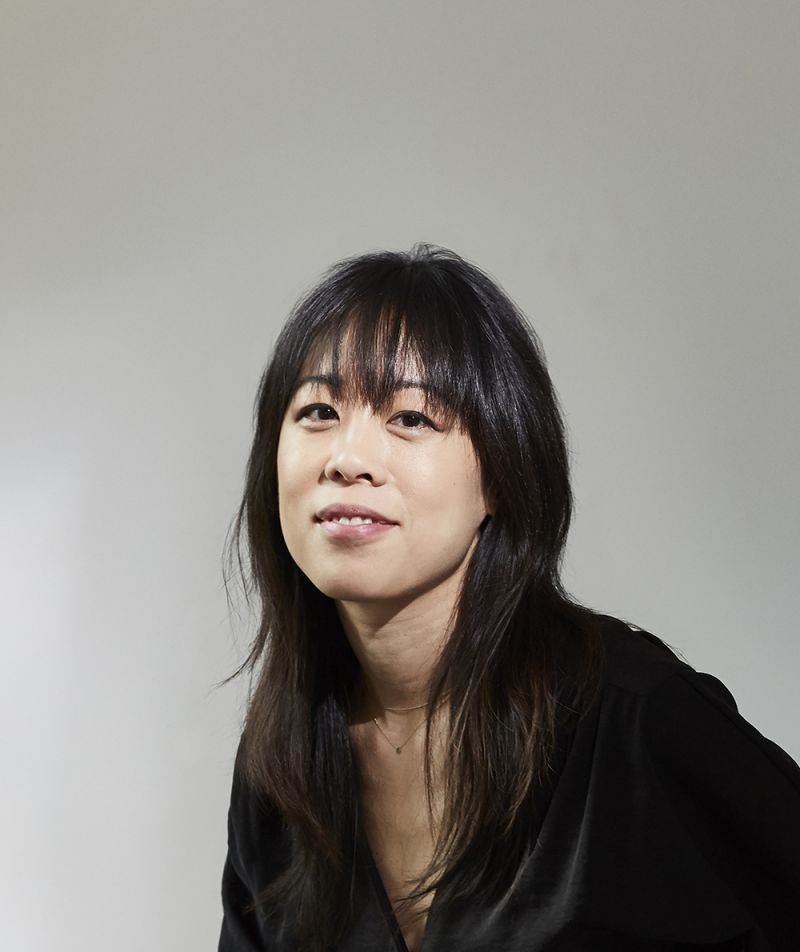 Color photo of an Asian woman with dark hair and top against a light gray backdrop