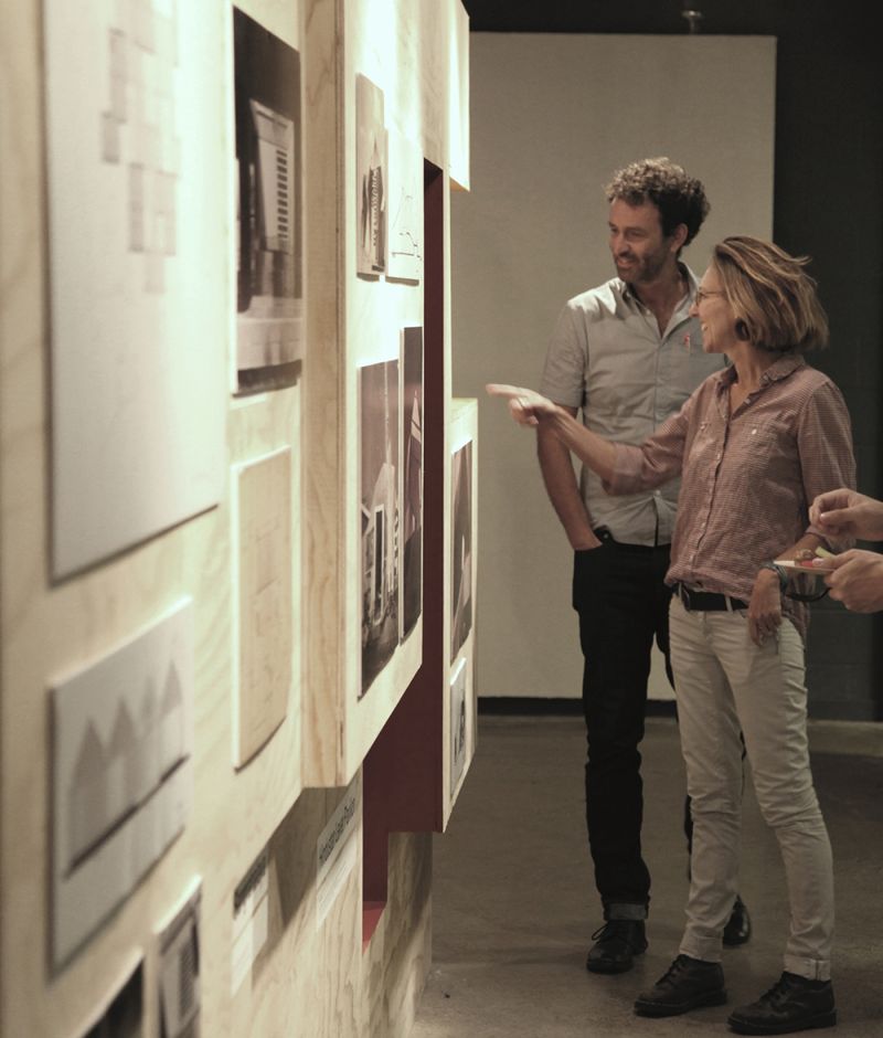 Color photograph of Mireille Roddier and Keith Mitnick looking at and pointing to a display of work on an exhibition wall
