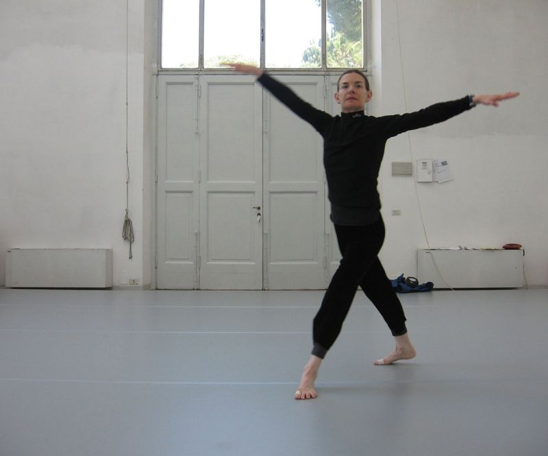 Color photograph of a light skinned woman in a dark bodysuit dancing in a white-walled studio with her arms and legs outstretched