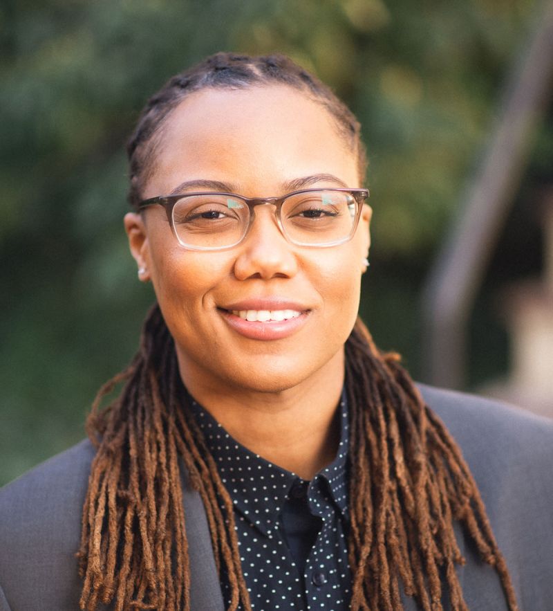 Color photograph of the head and shoulders of a dark skinned woman with long dreadlocks wearing glasses and smiling at the camera