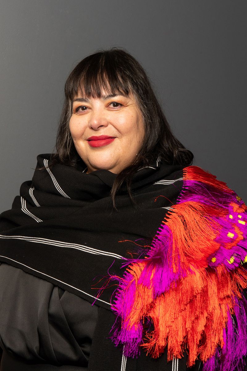 Color photograph of a light skinned woman with dark hair and bright red lipstick smiling at the camera; she wears a complex, multilayered garment in black, fucsia, and orange