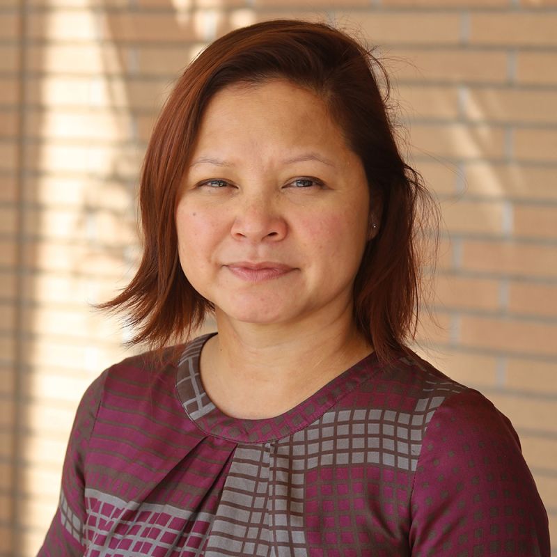 Color photograph of an Asian woman with auburn hair wearing a purple and gray gridded top; she stands in front of a brick wall and looks at the camera with a slight smile