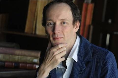 Color photograph of a light skinned man sitting in a study with one hand stroking his chin