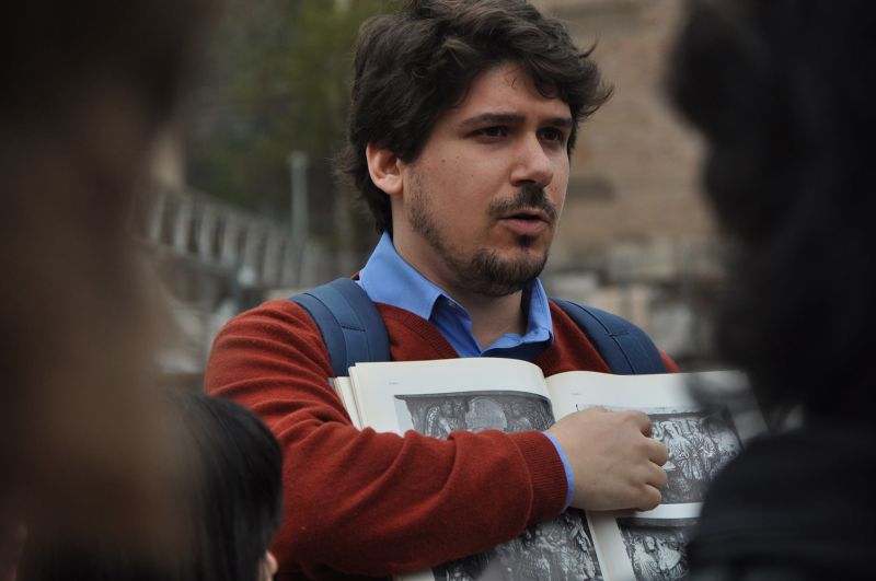 Color photograph of a light skinned man with dark hair holding a book and pointing to something in it