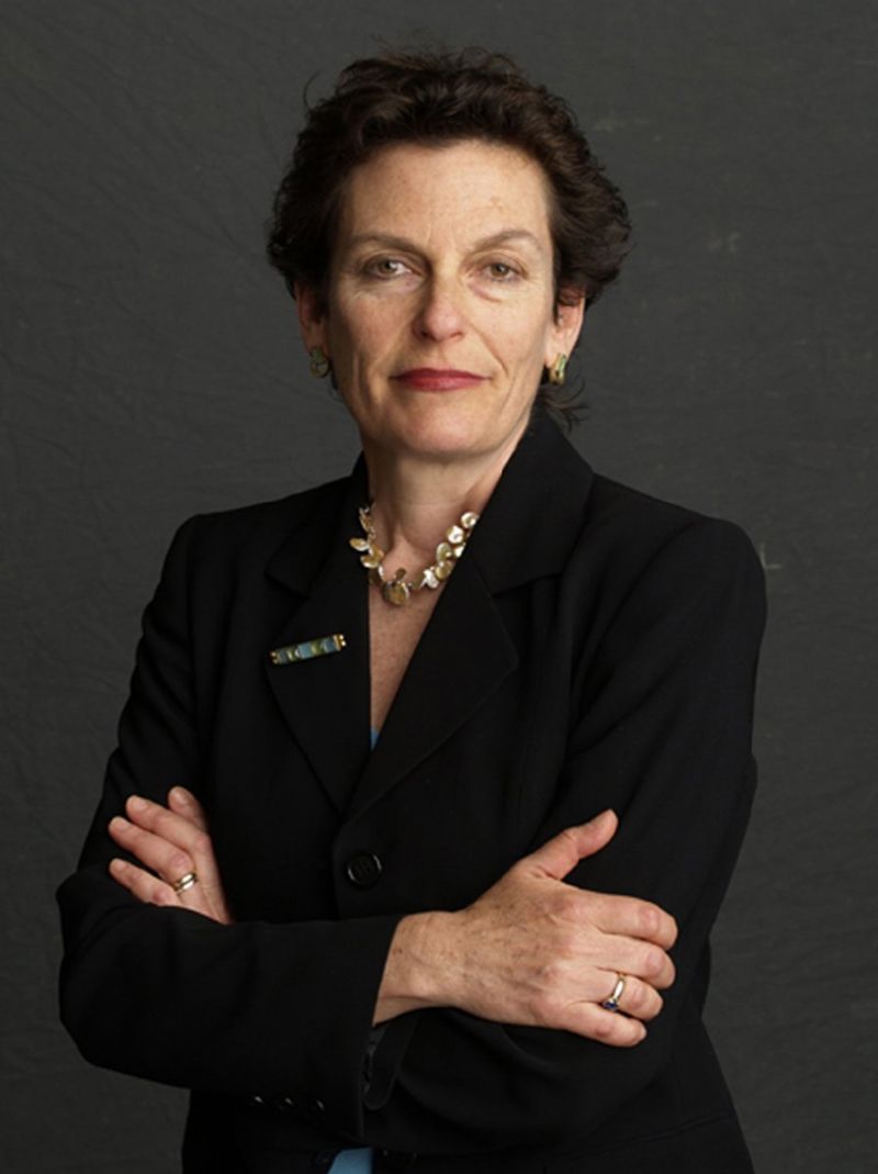 Color photograph of a light skinned woman seen from the waist up; she crosses her arms in front of her and looks at the camera with a serious expression