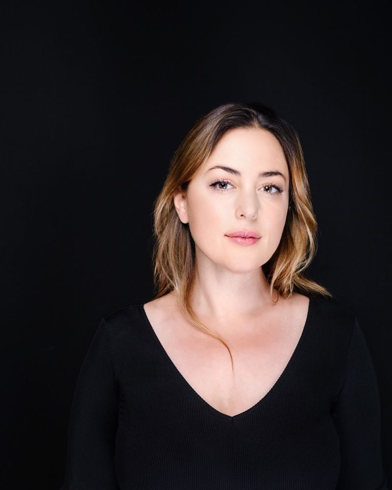 Color photograph of the head and torso of Paola Prestini, wearing a black top and sitting in front of a black backdrop