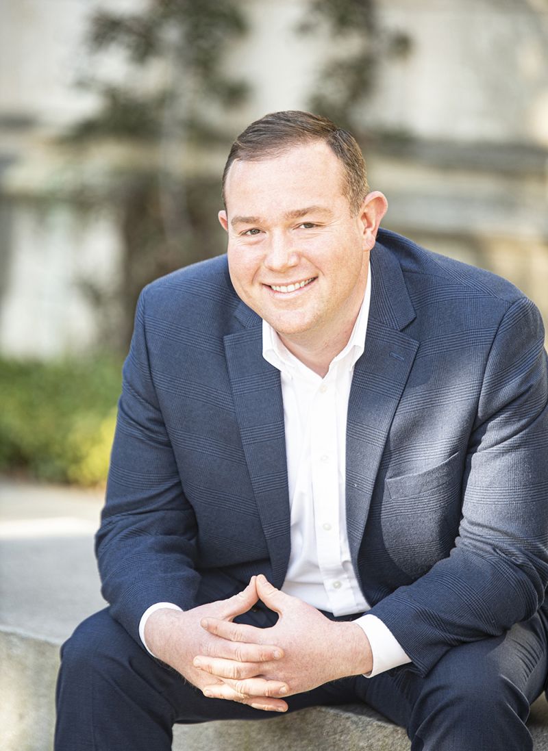 Color photograph of Randall Todd Pippenger wearing a suit and sitting outdoors on a concrete ledge