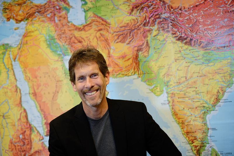 Color photograph of a light skinned man with brown hair smiling at the camera; he wears a dark blazer and is positioned in front of a large world map that spans from Egypt on the left to India on the right