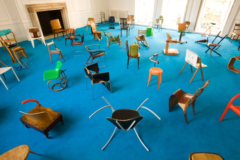 Color photograph of an indoor room with aqua blue carpet and a couple dozen assorted chairs