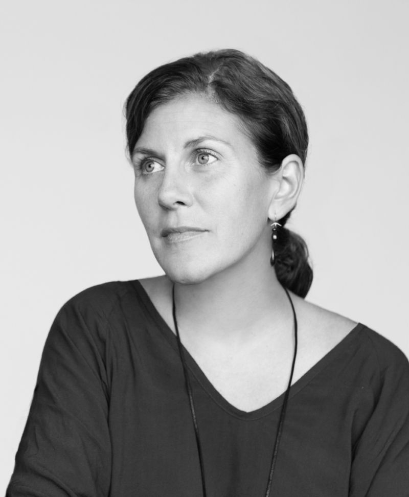 Black and white photograph of the head and torso of a light skinned woman with dark hair wearing a dark v neck top; she looks off camera to the left